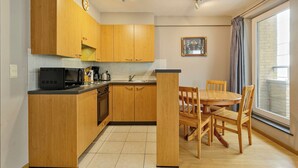 A compact kitchen with wooden cabinets, a microwave, and a dining area with a table and chairs. - Apartment in Blankenberge near the Beach (BLANKENBERGE)