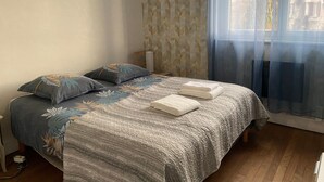 A bedroom with a bed, two pillows, a bedside table, a lamp, and a window with a view of houses. - LE ChicEthnic : maison de ville avec jardin, barbecue, jeux société (Besançon)