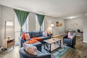 Una sala moderna con un sofá gris, una mesa de centro de madera y un sillón azul. Hay dos ventanas con cortinas verdes, una lámpara de pie y una mesita auxiliar con una planta.