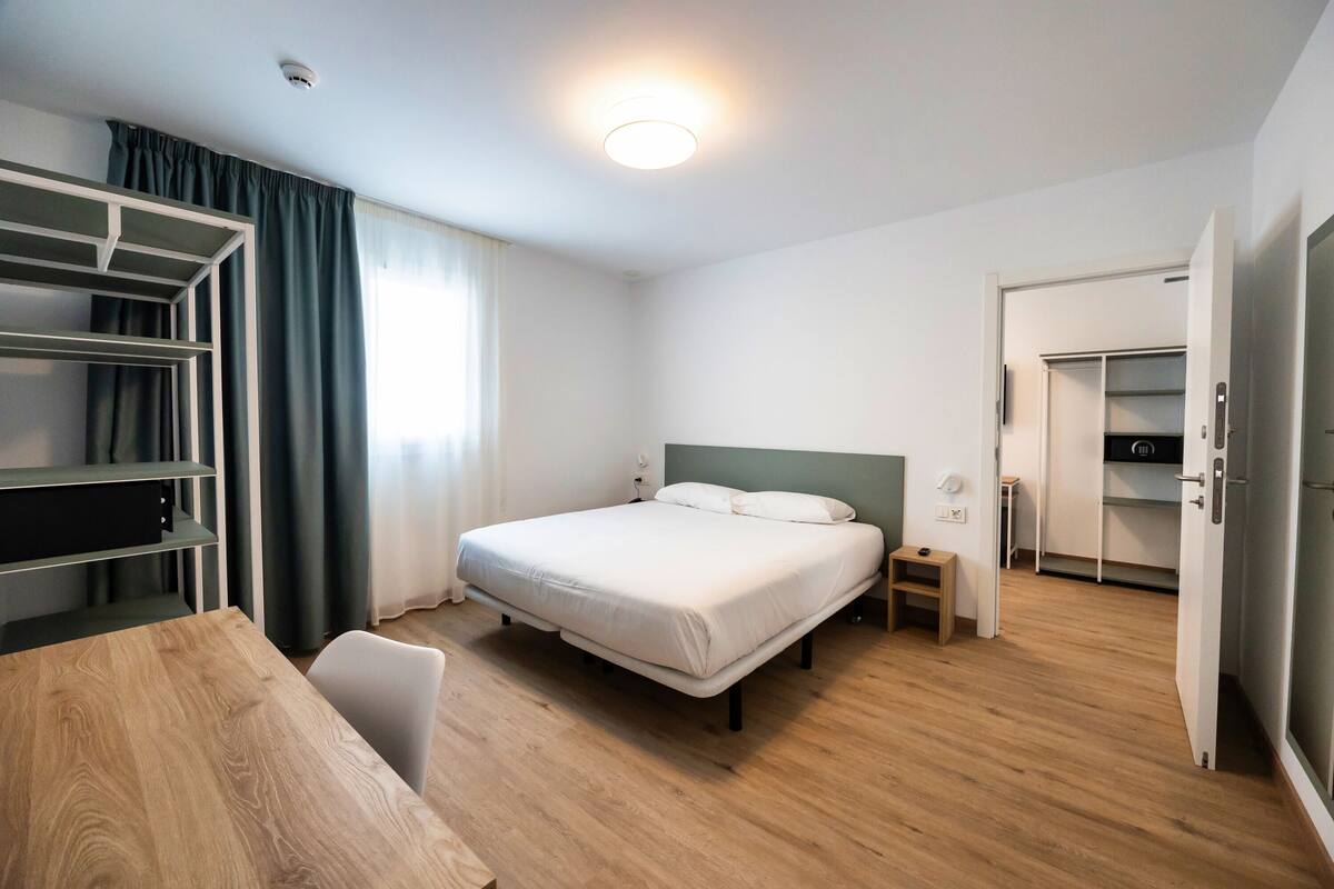 A hotel room with a bed, bedside table, wardrobe, and a desk with a chair.