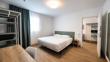 A hotel room with a bed, bedside table, wardrobe, and a desk with a chair.