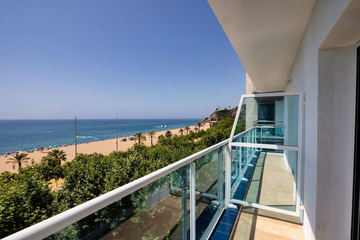 A balcony with a glass railing overlooking a beach with palm trees and the ocean.