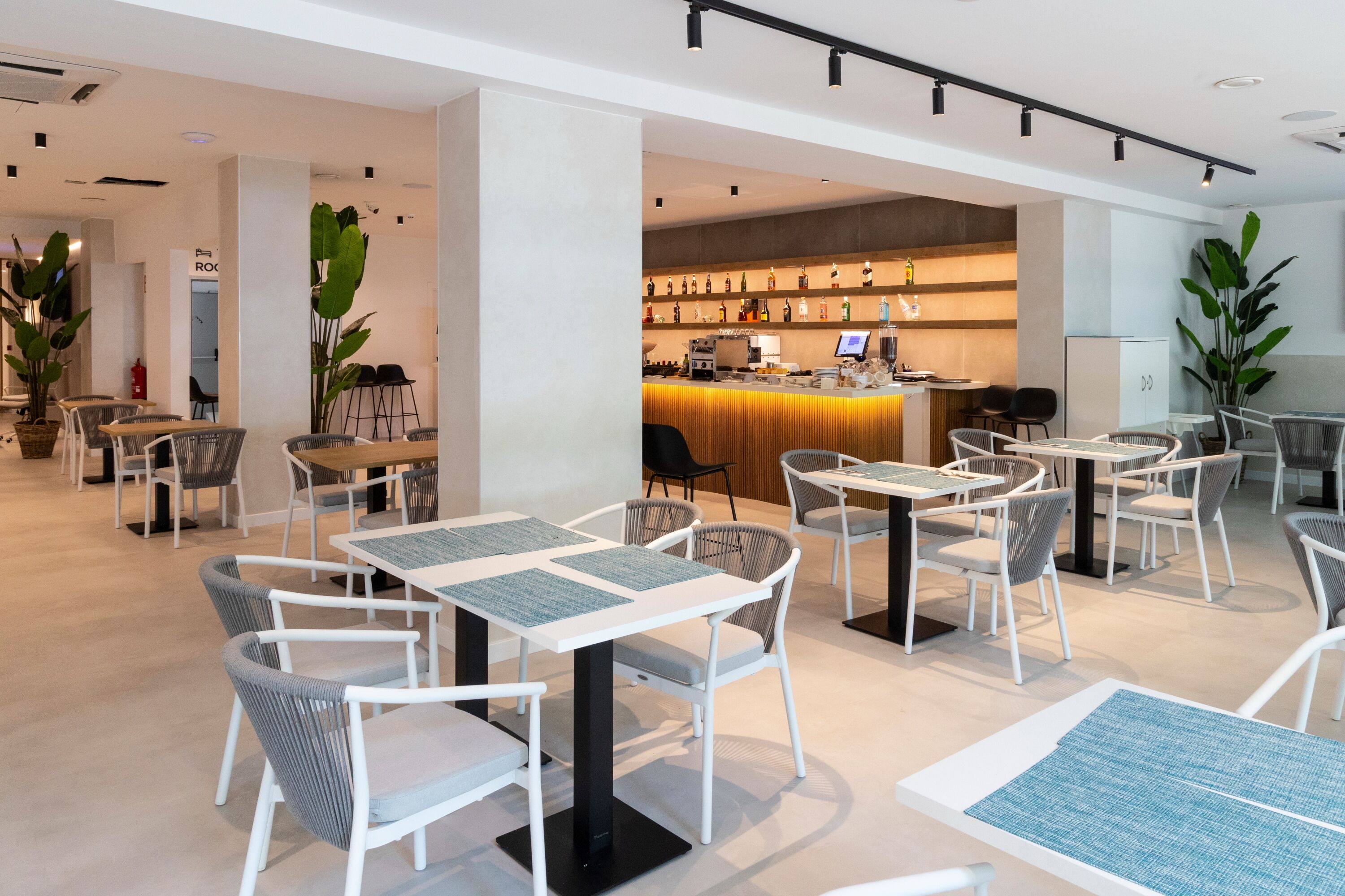 Un restaurante moderno con mesas y sillas blancas, una zona de bar con estantes llenos de botellas y grandes plantas en macetas.