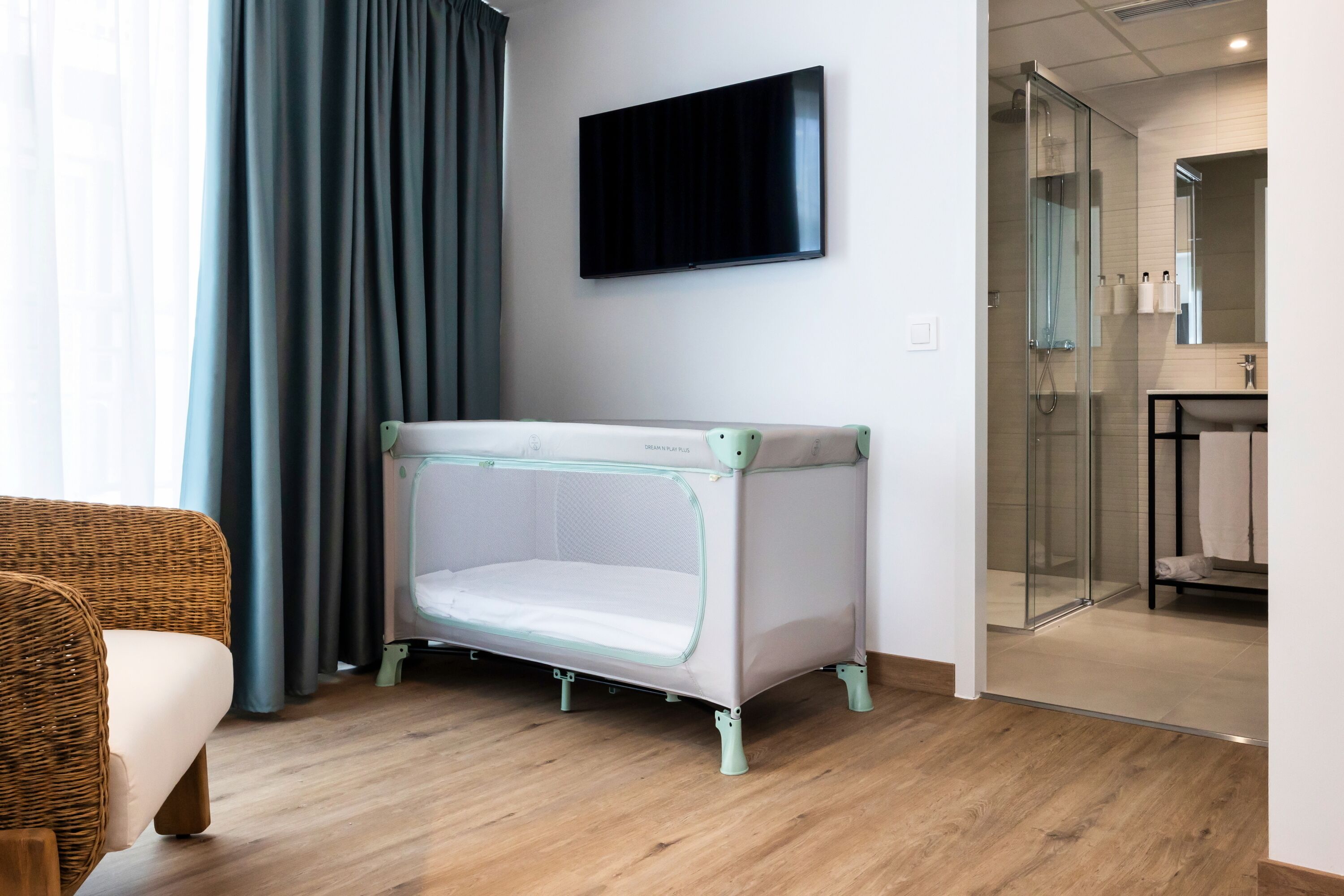 A modern hotel room with a baby crib, a television, and a glass-enclosed shower area.