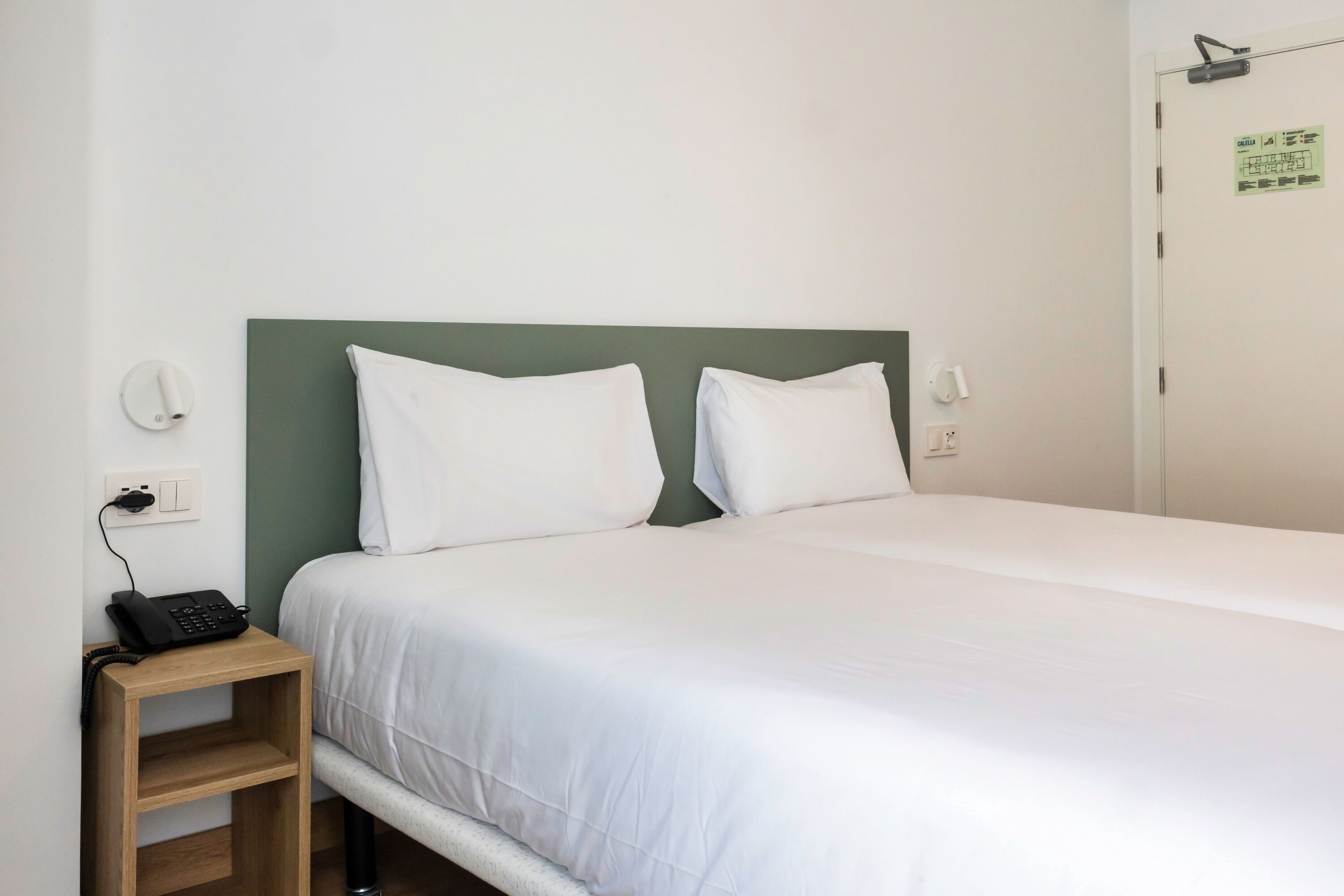 Una cama individual con ropa de cama blanca, una mesita de noche de madera con teléfono, y un cabecero verde.