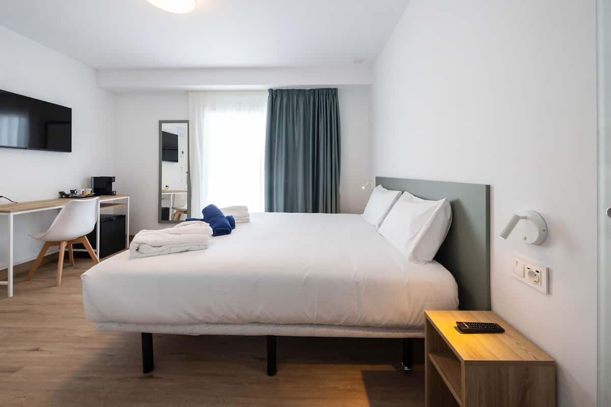A modern hotel room with a large bed, a desk with a chair, a TV mounted on the wall, and a window with curtains.