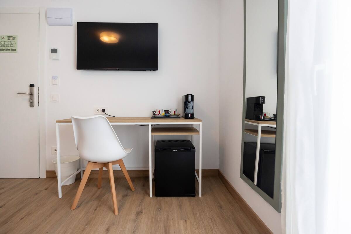 A small, modern room with a wooden desk, a white chair, a black mini-fridge, a television, and a large mirror.