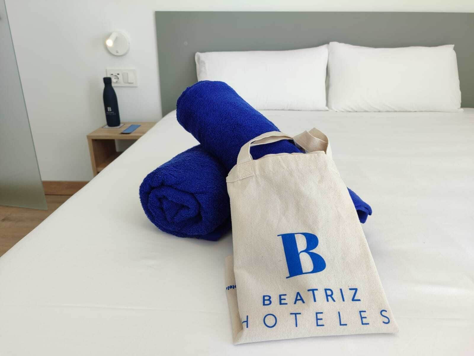 Una cama bien tendida con sábanas blancas, una toalla azul y una bolsa de tela color beige con el logotipo de Beatriz Hotels.