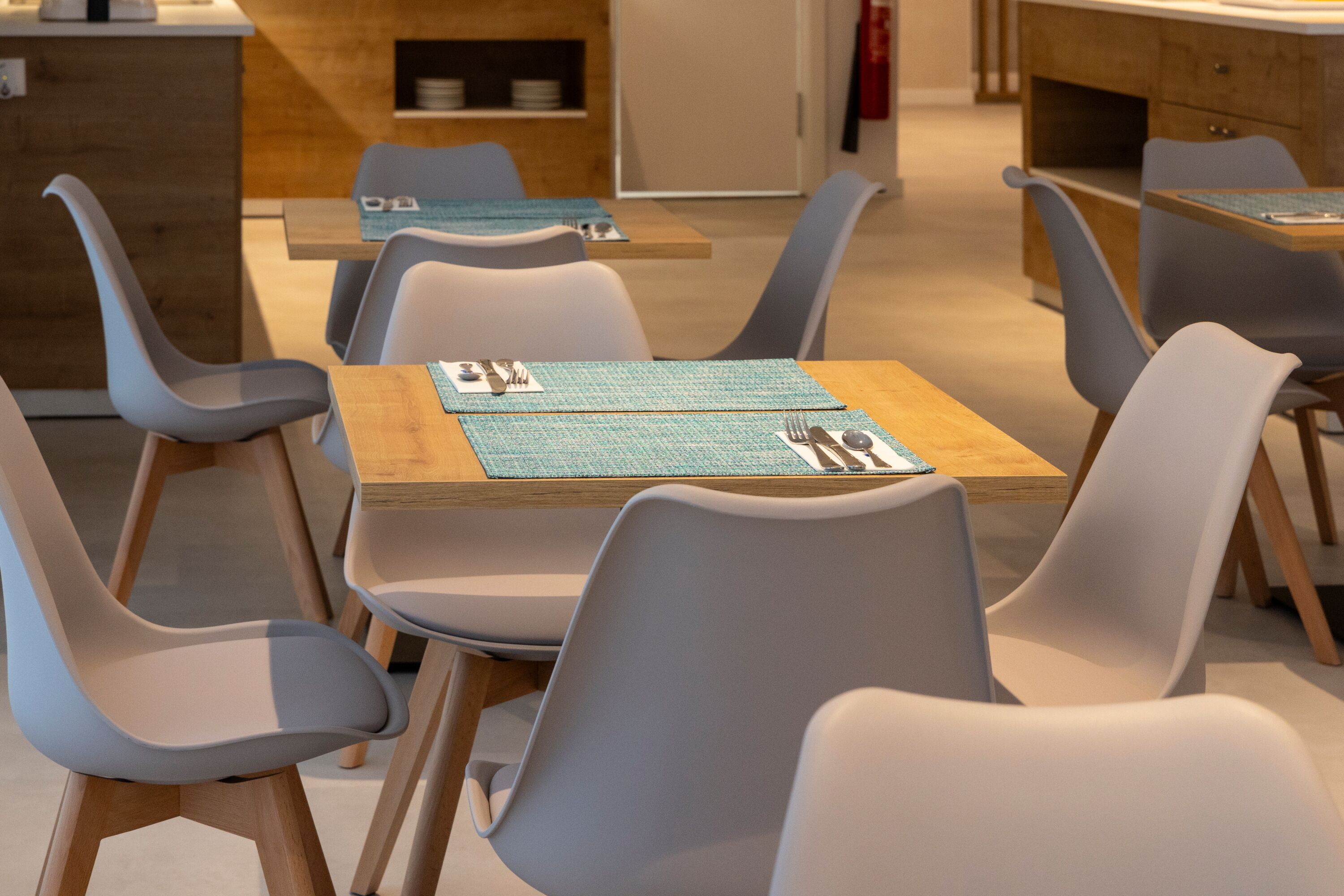 Un comedor moderno con mesas de madera y sillas grises, destacando una mesa central con posavasos y cubiertos.