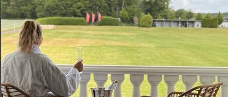 A person sitting on a balcony with wicker chairs, holding a glass of drink, overlooking a grassy field with flags and trees.