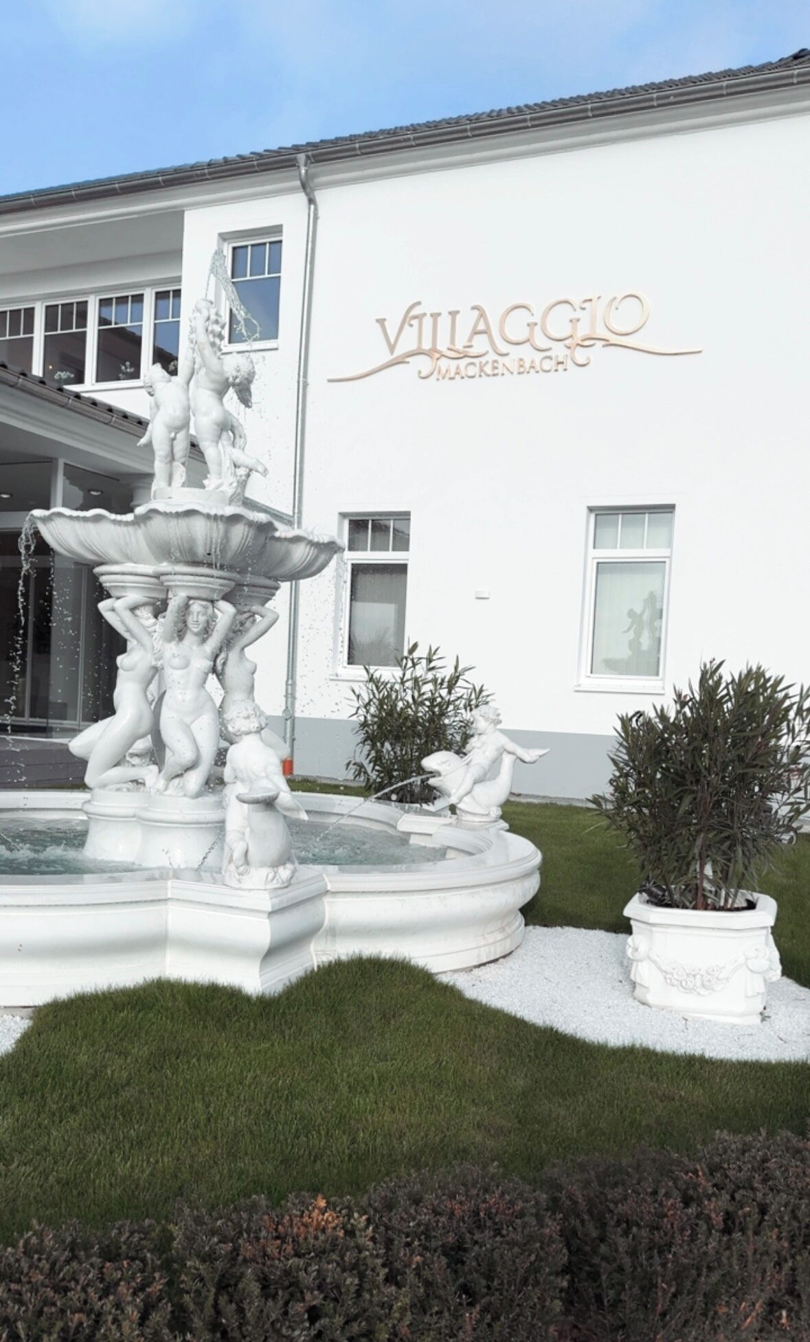 Un edificio bianco con un cartello che riporta "Villaggio Mackenbach". Di fronte, una fontana con statue e giochi d'acqua.