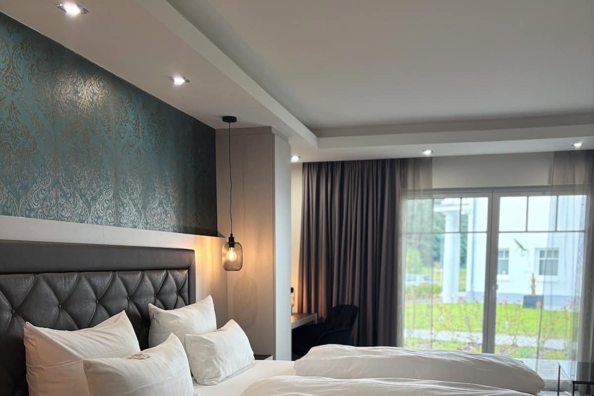 A modern hotel room with a large bed, a headboard, a bedside table, a lamp, and a view of the outdoors through a window.