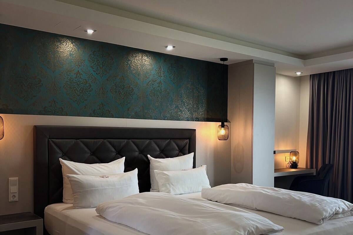 A modern hotel room with a large bed, a headboard with a tufted design, a wall-mounted light fixture, and a ceiling-mounted smoke detector.