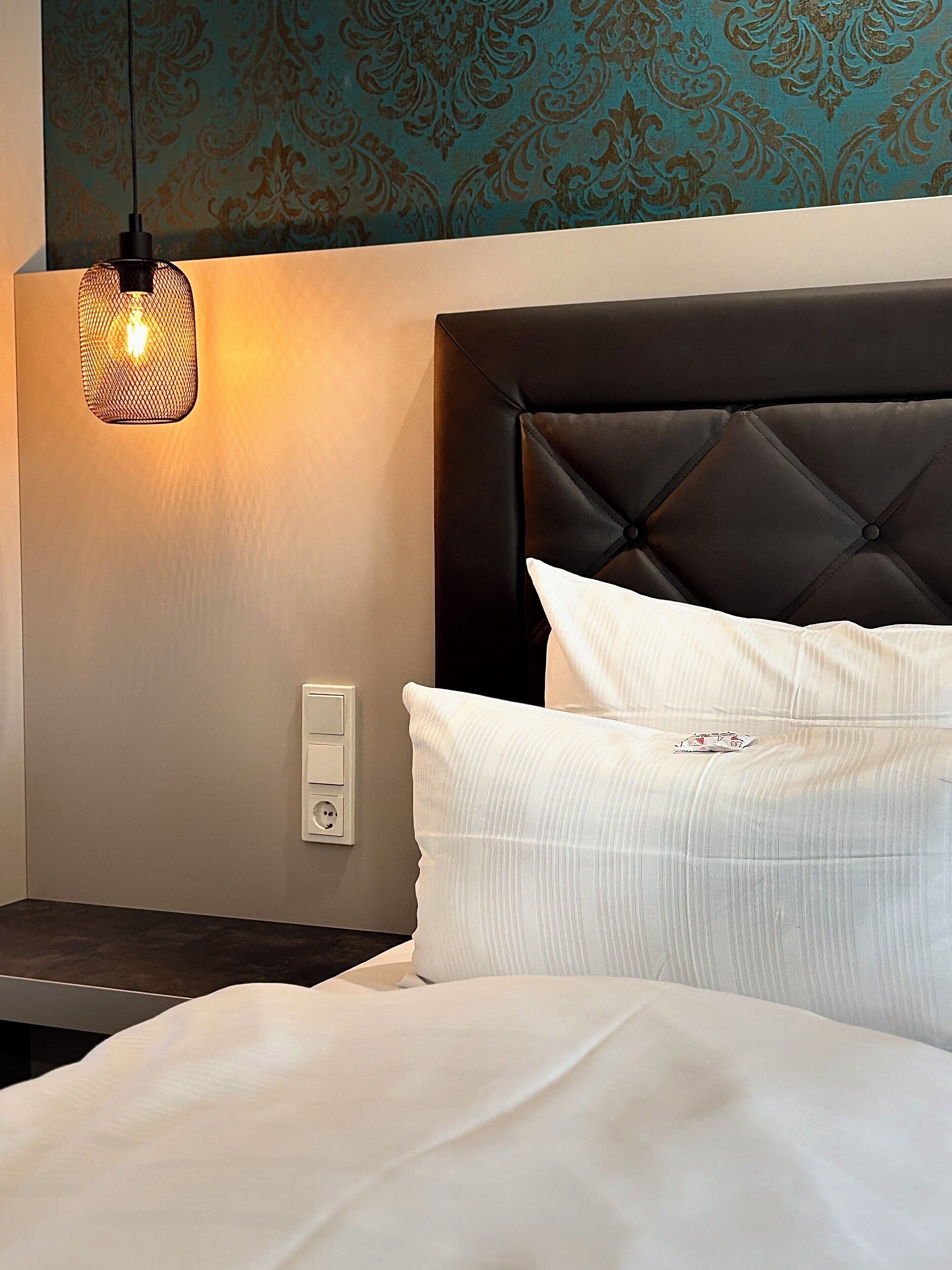 A hotel room with a tufted headboard, white bedding, a nightstand with a lamp, and patterned wallpaper.