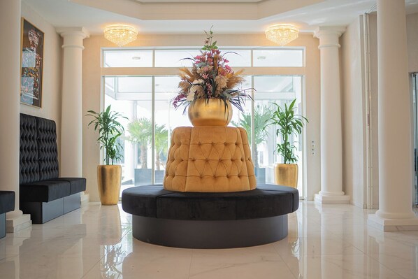 A lobby with a central round seating area, a large vase with flowers, and two potted plants. - Villaggio Hotel (Mackenbach)