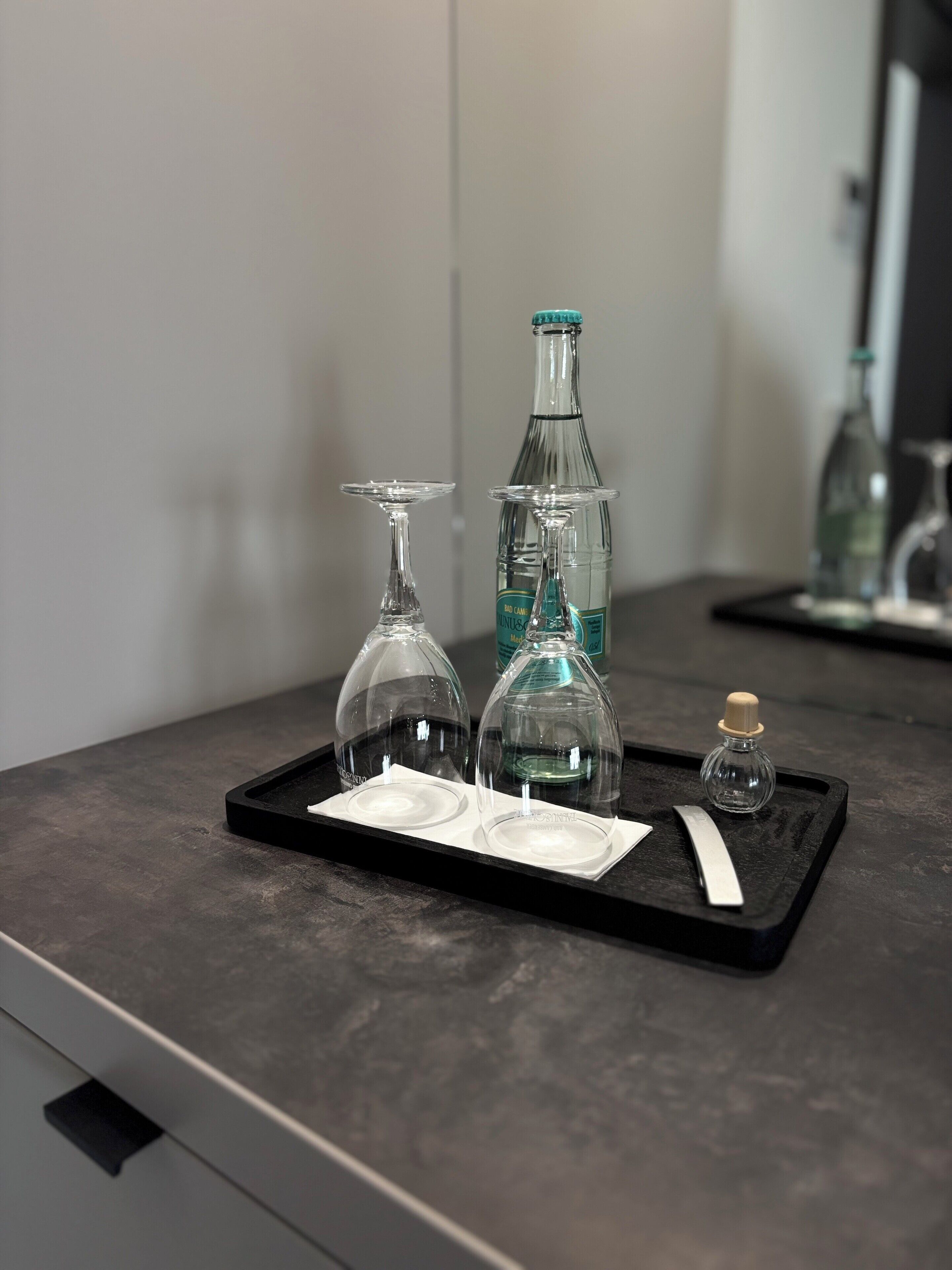 A tray with two wine glasses, a bottle, and a small bottle on a dark countertop.