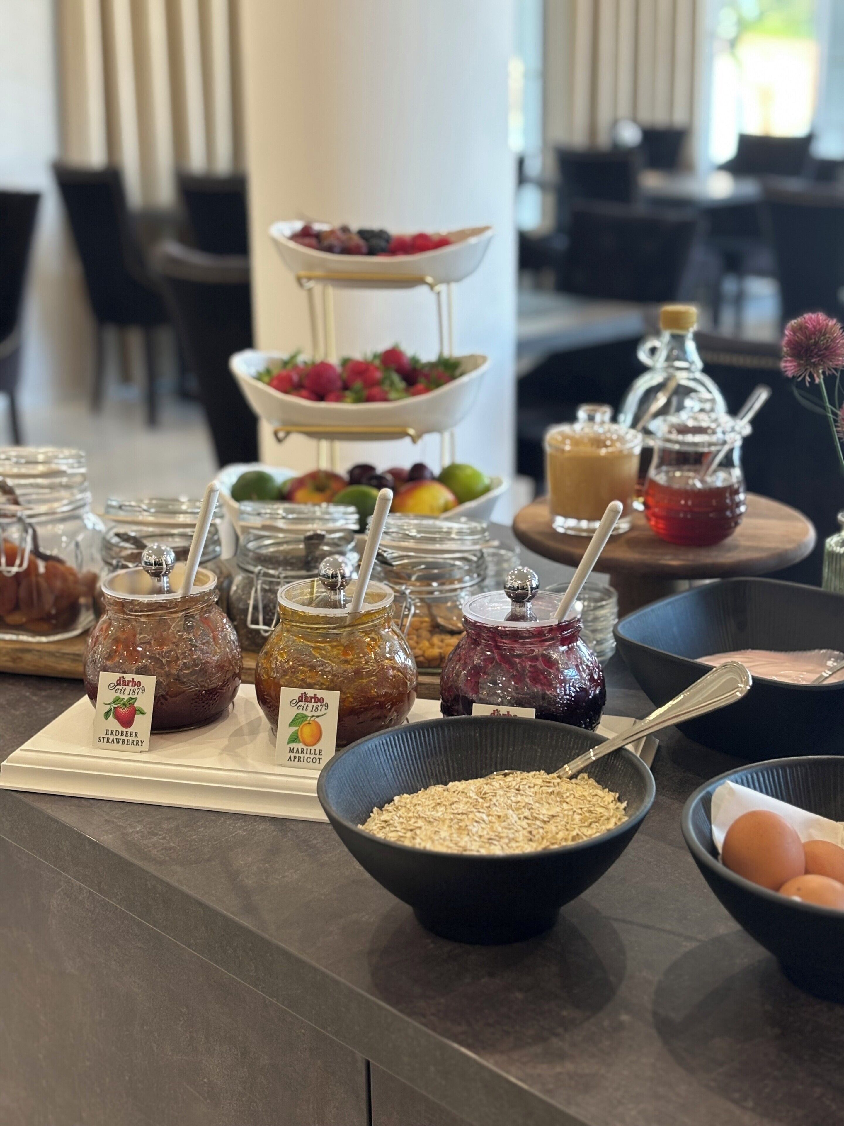 Una mesa de buffet con diversos alimentos, incluyendo frascos de mermelada, tazones de frutas y un bol de avena.
