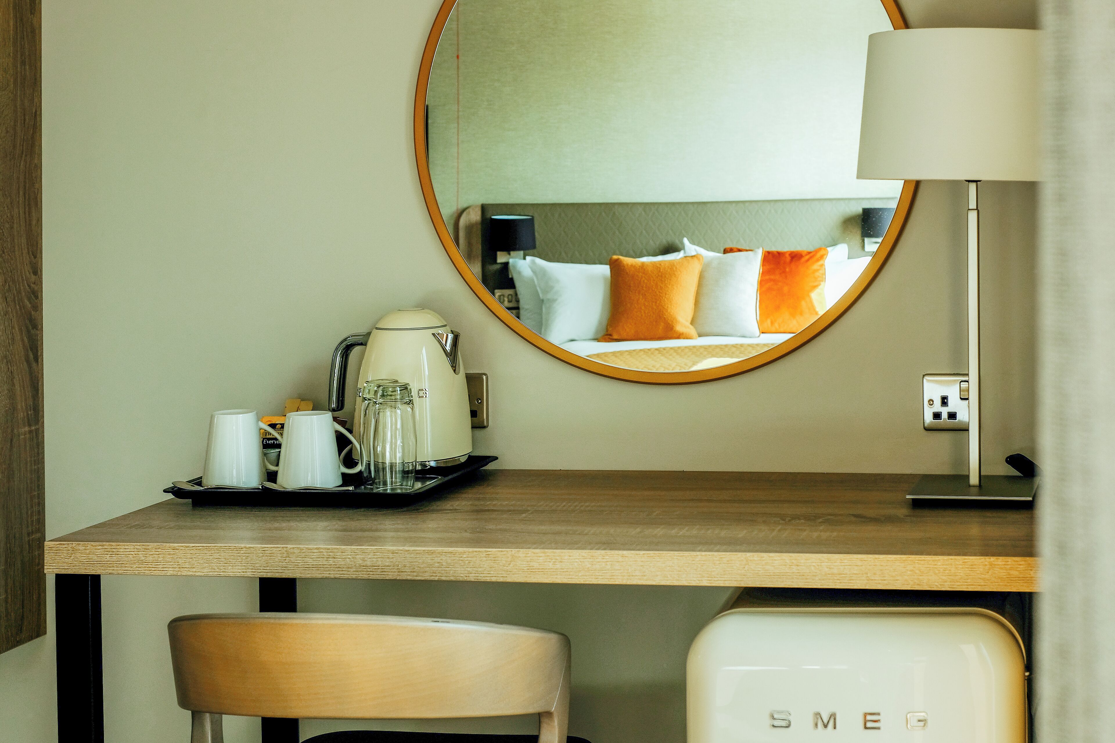 A hotel room with a wooden desk, a chair, a bed with orange and white pillows, a round mirror, a lamp, and a coffee maker.