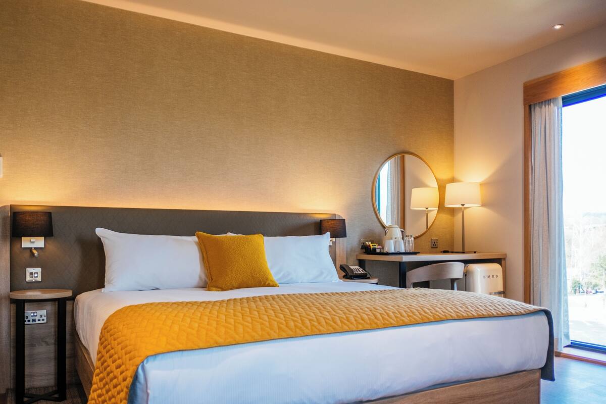 A hotel room with a large bed, a bedside table with a lamp, a desk with a chair, and a large window with curtains.