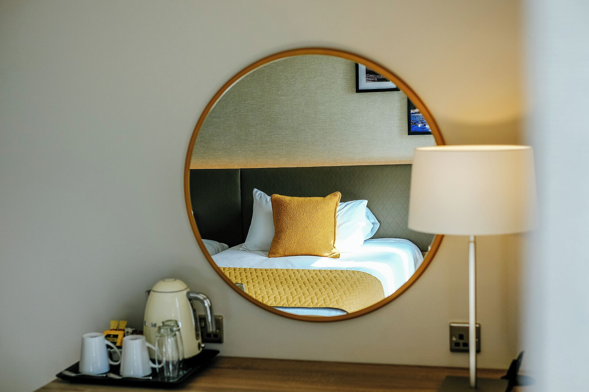 Una habitación de hotel con un espejo redondo que refleja una cama con almohadas blancas y mostaza, una mesita de noche con una lámpara y una bandeja con una tetera y tazas.