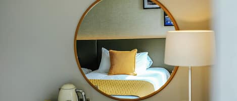Una habitación de hotel con un espejo redondo que refleja una cama con almohadas blancas y mostaza, una mesita de noche con una lámpara y una bandeja con una tetera y tazas.