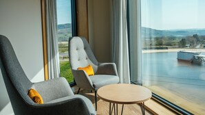 A modern room with two grey armchairs, a small round table, and large windows offering a view of a landscape with hills and a dam.