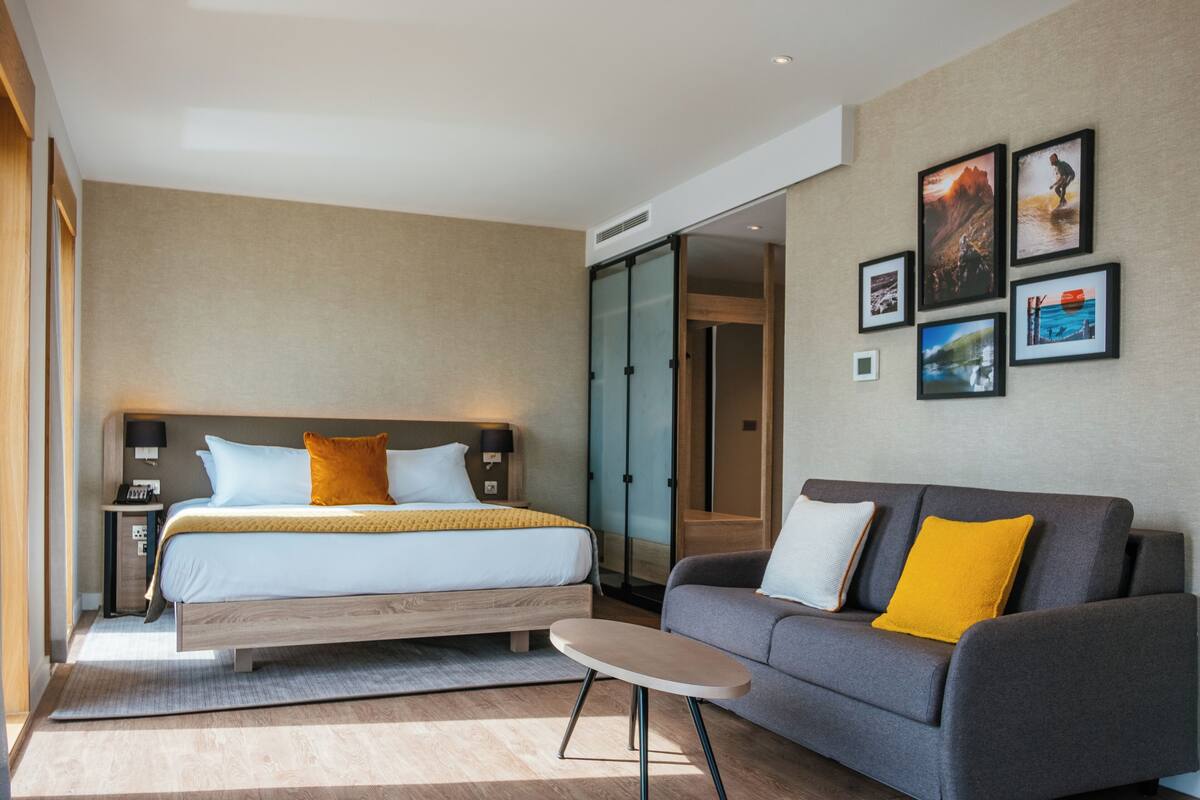 A modern hotel room with a large bed, a sofa, a small table, and a wall with framed pictures.