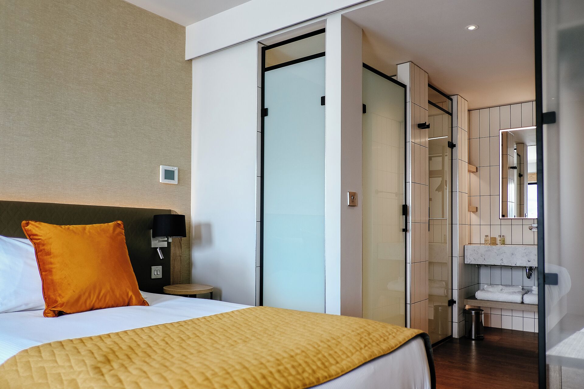 A hotel room with a bed, a nightstand, a lamp, a sliding door, a bathroom with a mirror, and a trash bin.