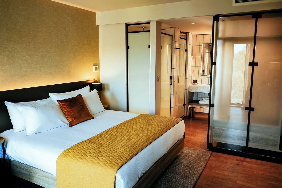 A hotel room with a bed, a bedside table, a lamp, a wardrobe, and a bathroom visible through a sliding door.