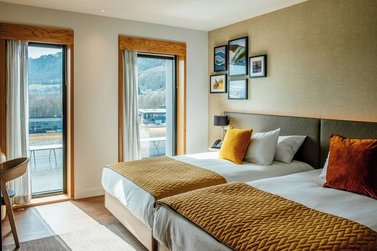 A hotel room with two beds, a view of mountains through the window, and framed pictures on the wall.