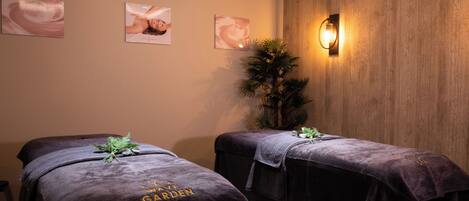 A spa room with two massage tables covered in dark towels, a wooden wall, and a wall-mounted light fixture.