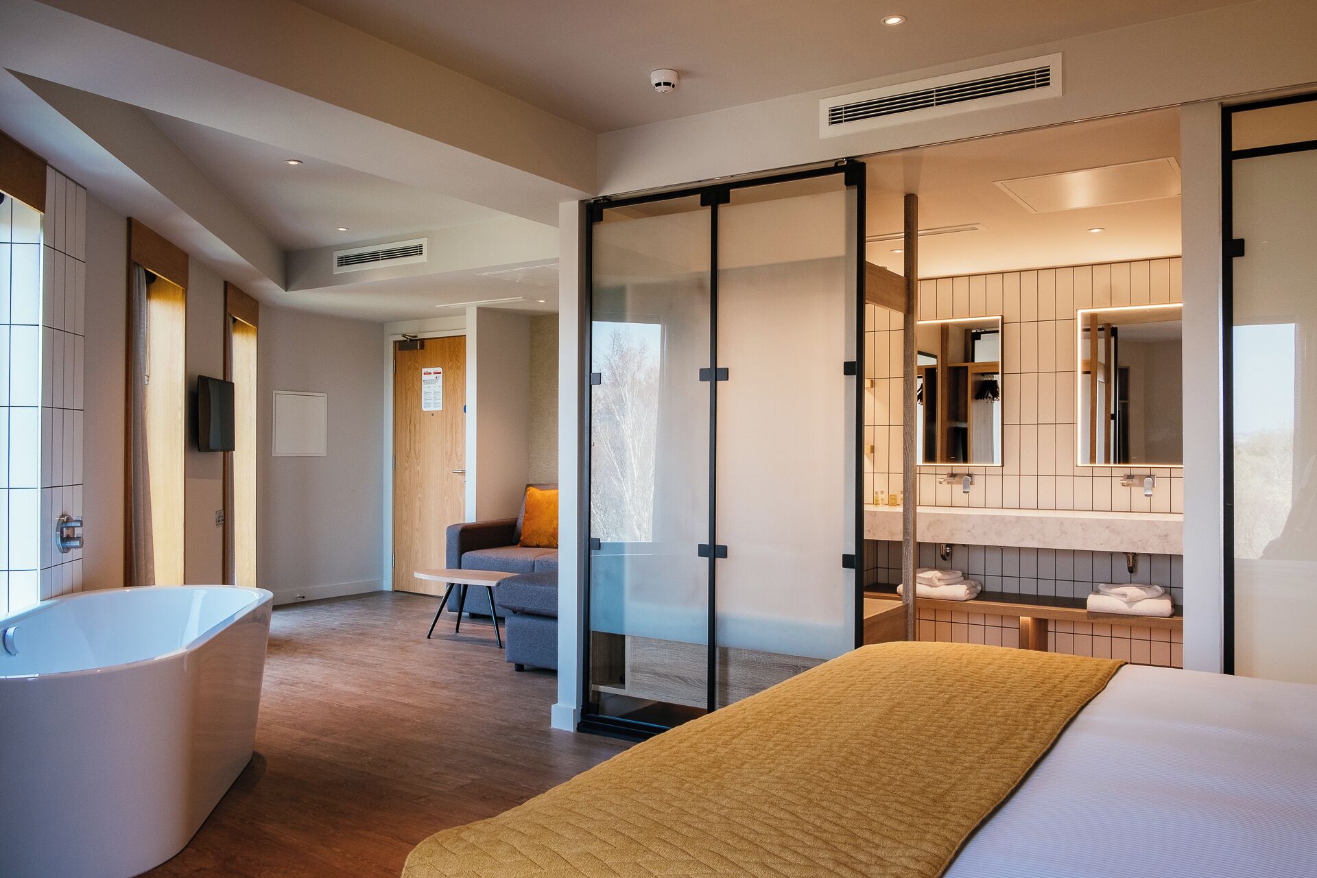 A modern hotel room with a large bed, a bathtub, a seating area with a chair, and a glass partition separating the bathroom from the bedroom.