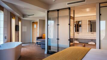 A modern hotel room with a large bed, a bathtub, a seating area with a chair, and a glass partition separating the bathroom from the bedroom.