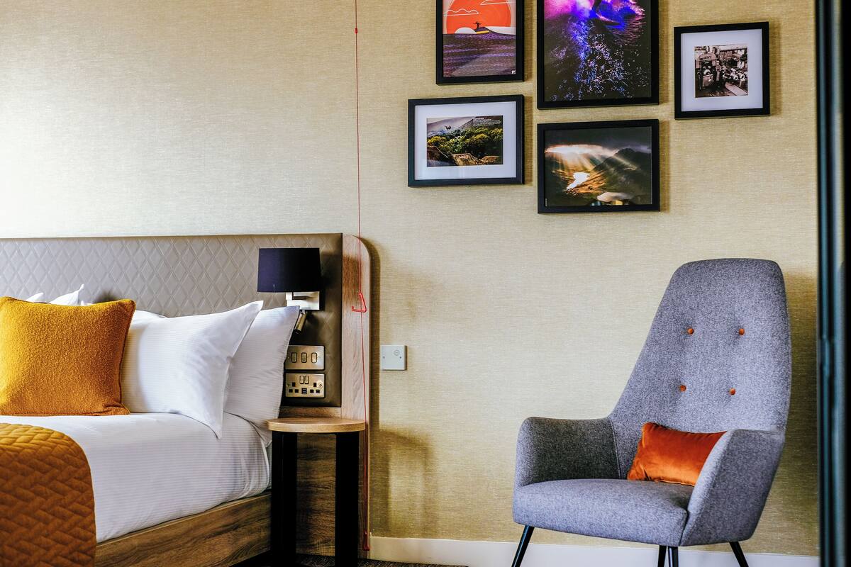 A hotel room with a bed, a bedside table with a lamp, a chair, and framed pictures on the wall.