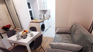 A compact living space with a dining table set for two, a grey sofa, a washing machine, and a small kitchen area. - Apartment Mama H (Dubrovnik)
