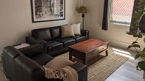 Ein Wohnzimmer mit einer schwarzen Ledersofa, einem Holztisch, einem gerahmten Bild an der Wand, einer Stehlampe und einem Fenster mit Blick auf eine Ziegelwand.