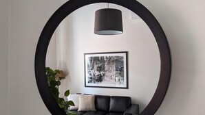 Ein runder Spiegel mit schwarzem Rahmen, ein schwarzes Sofa, ein weißes Kissen, eine Topfpflanze und eine schwarze Pendelleuchte.