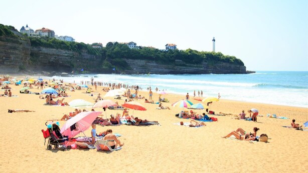 A crowded beach with people sunbathing and swimming, umbrellas, and a lighthouse in the background.