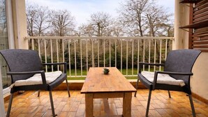 A balcony with two wicker chairs and a wooden table, overlooking a garden with leafless trees.