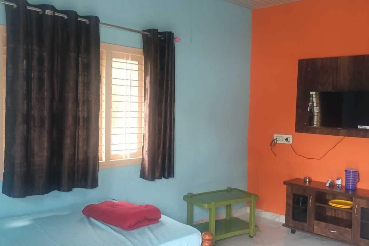 A room with a bed, a small table, a TV mounted on the wall, and a window with curtains.
