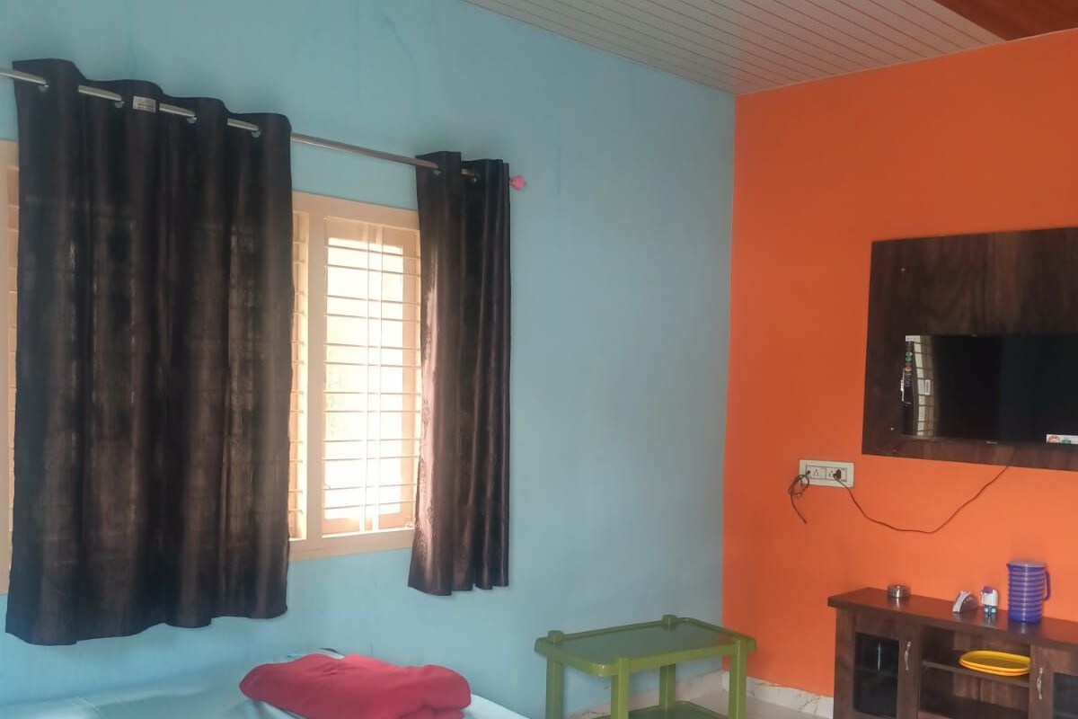 A room with a bed, a small table, a TV mounted on the wall, and a window with curtains.