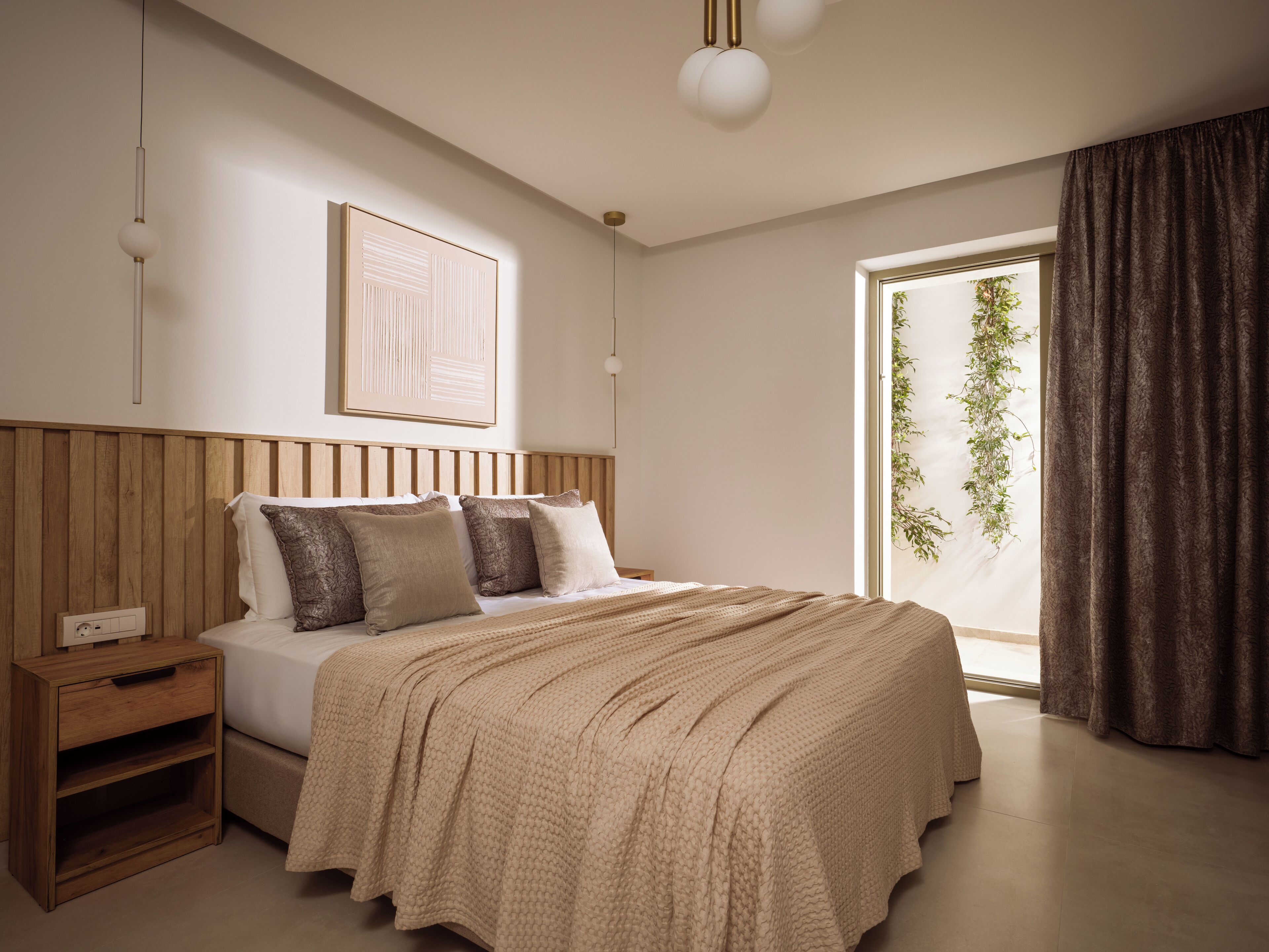 A bedroom with a wooden headboard, a bed with a beige blanket, a bedside table, and a large window with a view of greenery.