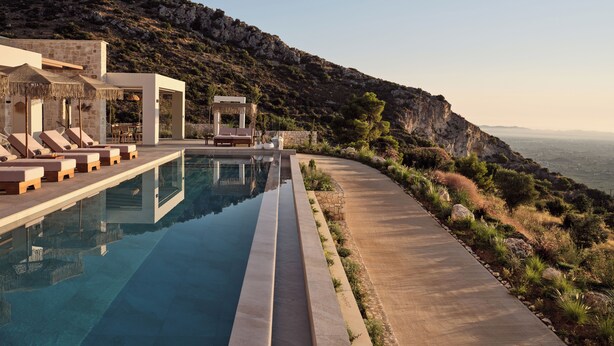 A modern pool area with lounge chairs and a thatched umbrella, surrounded by a stone wall and overlooking a mountainous landscape.