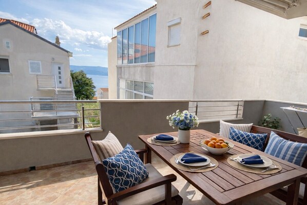 A terrace with a wooden table set for two, a bowl of fruit, and a vase of flowers.