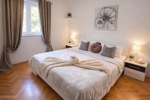 A bedroom with a large bed, two bedside tables, a window with curtains, a wall-mounted artwork, and a wooden floor.