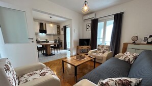 Ein modernes Wohnzimmer mit einer Couch, Sesseln und einem Couchtisch. Ein Fernseher ist vorhanden sowie ein kleiner Tisch mit einer Globus und Dekorationsgegenständen. Im Hintergrund ist eine Küche zu sehen.