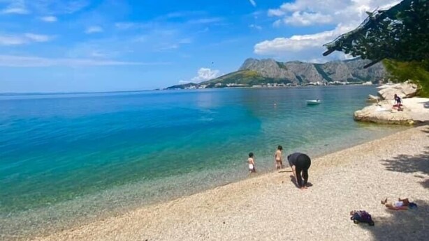 Ein Strand mit klarem türkisblauem Wasser, einem Felsen mit Menschen und einem Berg im Hintergrund.