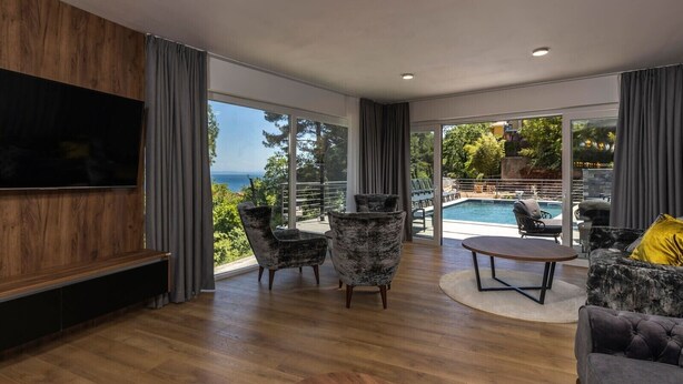A modern living room with a large TV, two armchairs, a coffee table, and a sofa. The room has large windows offering a view of a pool and the sea. - NEW! Villa with 6 Bedrooms and Heated Pool (Moscenicka Draga)