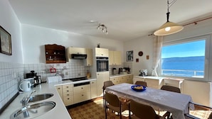Une cuisine compacte avec des meubles blancs, une cuisinière et un évier. On y trouve également un coin repas avec une table et des chaises, ainsi qu’une fenêtre donnant sur la mer.