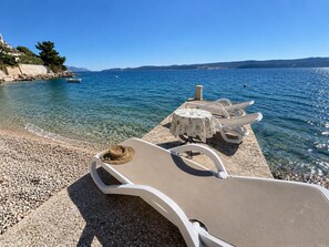 A clear beach with lounge chairs and a table, a rocky shoreline, and calm sea. - Azure Apartments By The Sea with 3 Bedrooms (Omis)